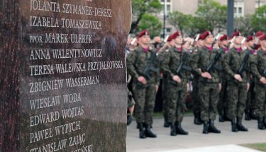 Na pierwszym planie fragment pomnika ofiar katastrofy smoleńskiej, w tle żołnierze wstawieni w szeregu