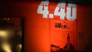 Ściana podświetlona na pomarańczowo, na niej wyświetlona godzina 4.04, niżej plakat z napisem: 1 września 1939 roku, Wieluń, pamiętamy